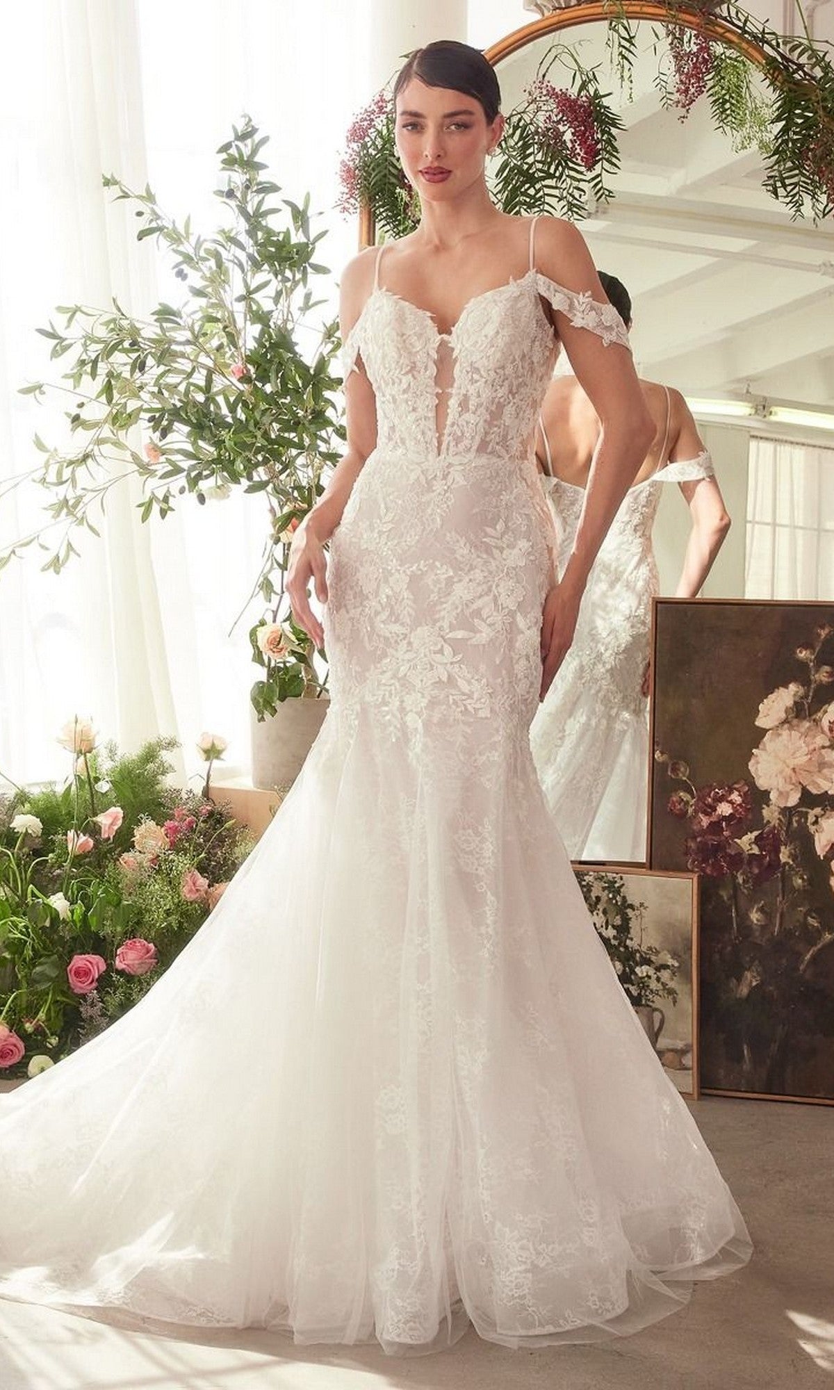 A woman stands indoors in the Andrea and Leo WL051 long wedding dress—a white lace off-shoulder mermaid gown with a long train and layered tulle skirt—surrounded by flowers and mirrors.